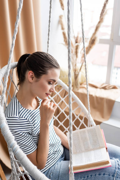 Selective focus of young woman reading book in hanging armchair at home  - Photo, image