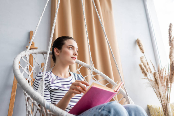 Selective focus of young woman holding book while sitting in hanging armchair  - Foto, immagini