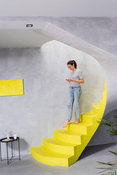 Selective focus of barefoot woman with smartphone walking on stairs at home  - Fotó, kép