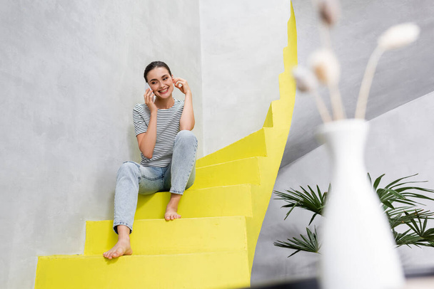 Foco seletivo da jovem mulher falando no smartphone enquanto está sentada nas escadas em casa  - Foto, Imagem