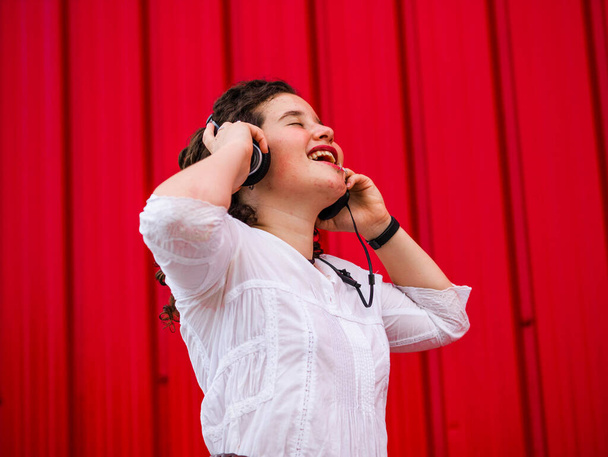 Молода кавказька жінка з радістю слухає розмови навушниками біля червоної стіни. - Фото, зображення