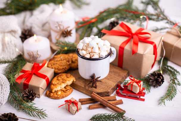 Enamelled cup of hot cocoa or coffee with marshmallows and cookies. Around the tree branches, gifts and burning candles. Christmas mood. Postcard or winter background. - Photo, Image