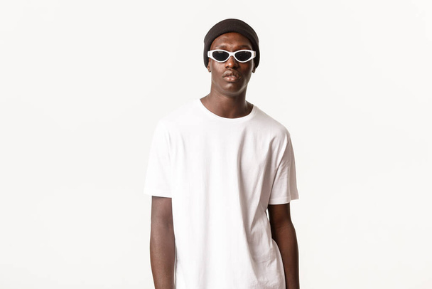 Portrait of stylish and cool african-american young guy in beanie and sunglasses, looking confident and sassy, standing over white background - Fotó, kép