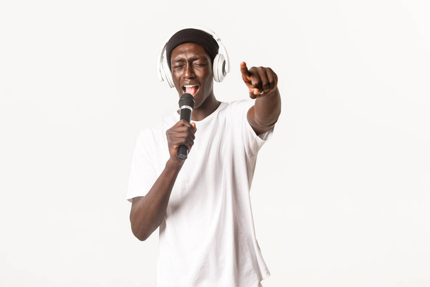 Portrait of carefree and cool african-american young man, performing in karaoke, singing into microphone while wearing headphones, white background - Foto, afbeelding
