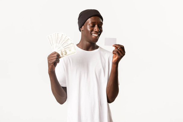 Şanslı, heyecanlı, Afro-Amerikalı genç bir adamın portresi. Kredi kartında mutlu görünüyor, elinde para, beyaz bir geçmişi var. - Fotoğraf, Görsel