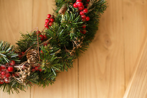 Kerstkrans opknoping op rustieke houten deur in huis.Traditionele kerstkrans met rode bessen en ornamenten, dennenappels en kaneel op houten achtergrond, vakantie decor. - Foto, afbeelding