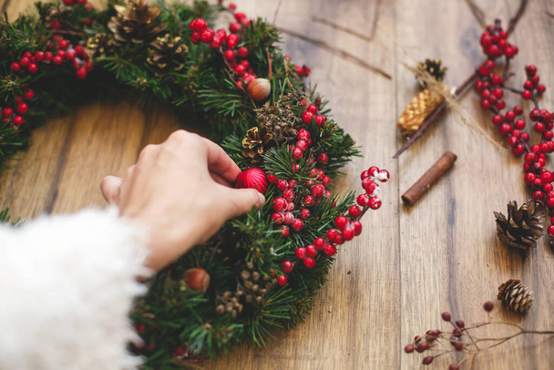 Kırsal ahşap arka planda geleneksel Noel çelengi yapan kişi. Çiçekçi el süsü, pamuk, kırmızı böğürtlen ve çam kozalaklarıyla Noel çelengi süslüyor. Tatil atölyesi - Fotoğraf, Görsel