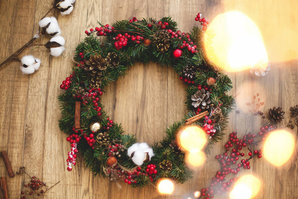 木製の素朴な背景フラットレイアウト上のクリスマスリース。赤い果実と装飾品、松のコーンと木の綿と伝統的なクリスマスの花輪。休日ワークショップのための装飾 - 写真・画像
