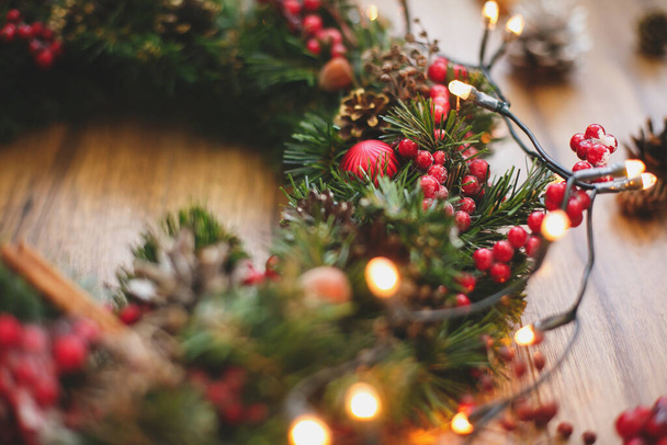 Weihnachtskranz-Dekorationen auf rustikalem Holz in Lichterketten. Rote Beeren, Schmuck und Tannenzapfen auf dem traditionellen Adventskranz, Weihnachtswerkstatt. Raum für Text - Foto, Bild