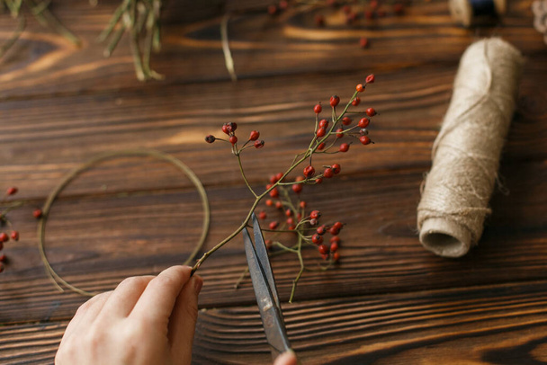 Making christmas wreath on rustic wooden table, holiday advent. Florist making simple christmas wreath with red berries, holding scissors. Festive workshop - Foto, Imagen