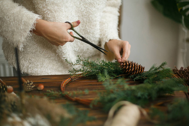 Изготовление деревенского рождественского венка, рождественское пришествие. Руки держат зеленые кедровые ветви на деревенском деревянном фоне. Сезонная праздничная мастерская или праздничные украшения дома - Фото, изображение