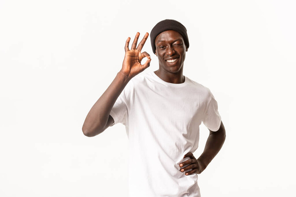 Portret van zelfverzekerde en vrolijke Afrikaans-Amerikaanse coole man in Beanie, verzekeren alle goede, knipperen in goedkeuring en tonen oke gebaar, garantie kwaliteit, witte achtergrond - Foto, afbeelding
