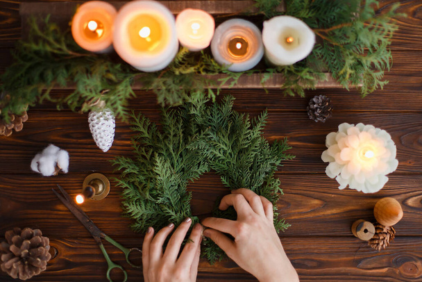 Rustic christmas wreath with candles, pine cones, scissors and ornaments on wooden table, flat lay. Female hands making simple stylish christmas wreath with fir branches, holiday advent - Photo, Image