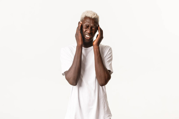 Retrato de un joven rubio afroamericano con dolor de cabeza severo, tocando la cabeza y haciendo muecas por el dolor, sufriendo migraña, de pie fondo blanco - Foto, Imagen