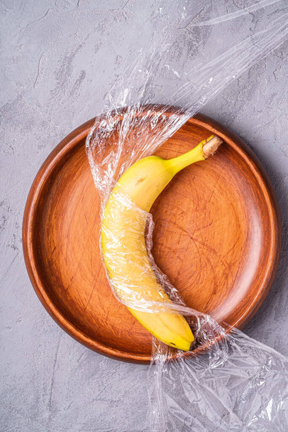 Bananenfruit verpakt in rekfolie plastic in houten plaat, steen beton achtergrond, creatieve lay-out, ecologie en milieu concept - Foto, afbeelding