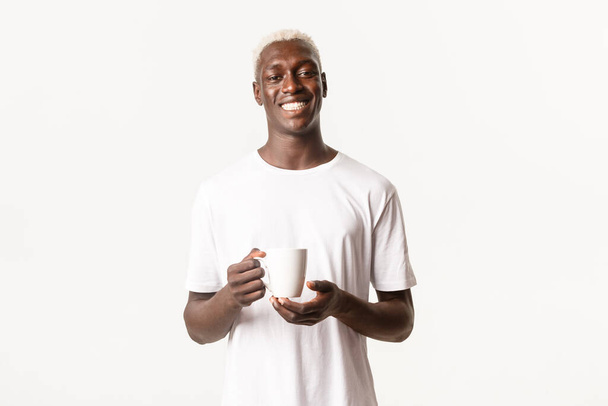 Porträt eines glücklich lächelnden afrikanisch-amerikanischen blonden Mannes, der zufrieden aussieht, Kaffee oder Tee trinkt und vor weißem Hintergrund steht - Foto, Bild