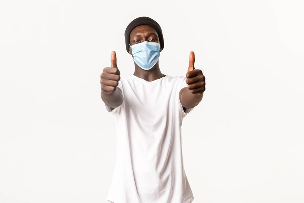 Portret van tevreden Afrikaans-Amerikaanse man in muts en medisch masker, met duimen omhoog in goedkeuring, staande witte achtergrond - Foto, afbeelding
