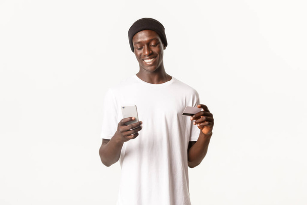 Retrato del guapo hombre afroamericano sonriente comprando en línea, con tarjeta de crédito y teléfono móvil, fondo blanco - Foto, imagen