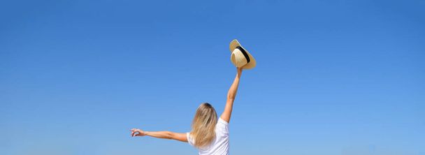 Mavi gökyüzüne el kaldırmış şapkalı bir kızın arka görüntüsü. - Fotoğraf, Görsel