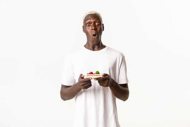 Portret van een verraste Afrikaans-Amerikaanse blonde man, zeg wow, kijkend verwonderd en met een stuk taart, staande witte achtergrond - Foto, afbeelding