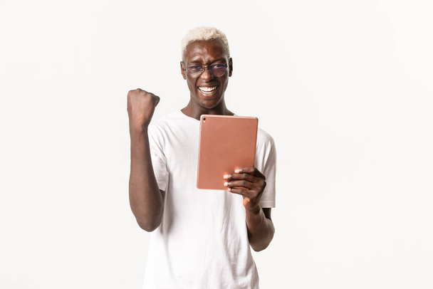 Porträt des glücklichen Gewinners afrikanisch-amerikanischer Typ, trägt eine Brille, hält ein digitales Tablet in der Hand und lässt die Faust jubeln, lächelt und triumphiert, erreicht das Ziel in App, weißer Hintergrund - Foto, Bild