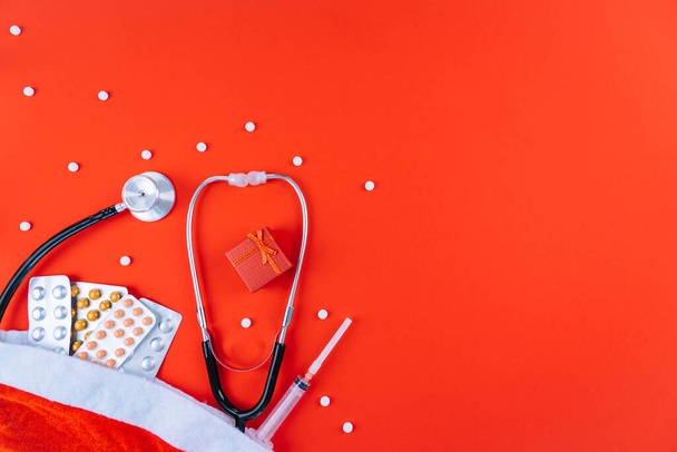 Geneeskunde kerst nieuwjaar concept banner tijdens coronavirus covid-19 tijd. Bovenaanzicht van medische pillen, wegwerp beschermend gezichtsmasker, geschenkdoos, stethoscoop in santa hoed op rode achtergrond. CopySpace - Foto, afbeelding