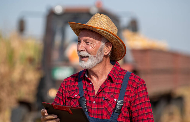 Starszy rolnik w słomianym kapeluszu trzymający notatnik i stojący przed ciągnikiem z przyczepą pełną kolby kukurydzy podczas zbiorów - Zdjęcie, obraz