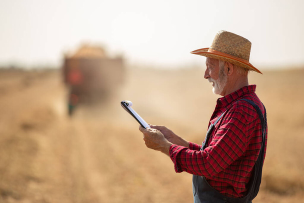 Ritratto di agricoltore anziano con cappello di paglia che guarda le note davanti al trattore con rimorchio che lavora nel campo di mais durante il raccolto - Foto, immagini