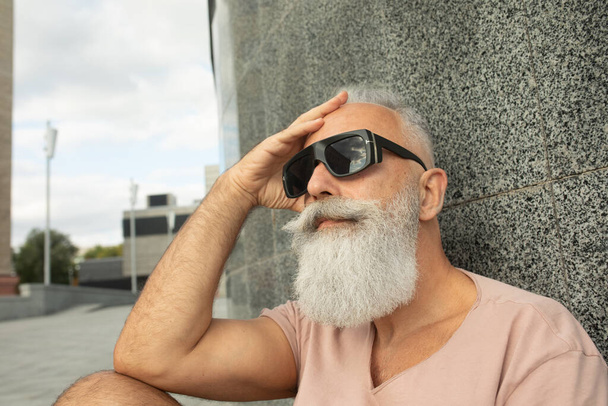 Close view of hipster mature man wearing sunglasses leaning on building facade in the city - Photo, image