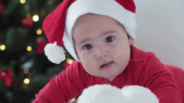 merry xmas and happy new year, infants, childhood, holidays concept - close-up 6 month old newborn baby in santa claus hat and red bodysuit on his tummy crawls with decorations balls on christmas tree - Filmagem, Vídeo