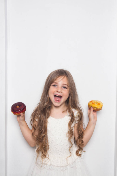 Маленькая счастливая милая дошкольница ест разноцветные пончики на белом фоне изолированы. Эмоциональный ребенок веселится со сладостями. Вкусная сладкая еда для детей. Бесплатное пространство для копирования текста макета  - Фото, изображение