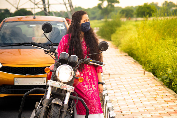 Nuori intialainen nainen punaisessa salwar kameez ja naamio nojaten pyörä oranssi auto taustalla ja feilds puolella tietä - Valokuva, kuva