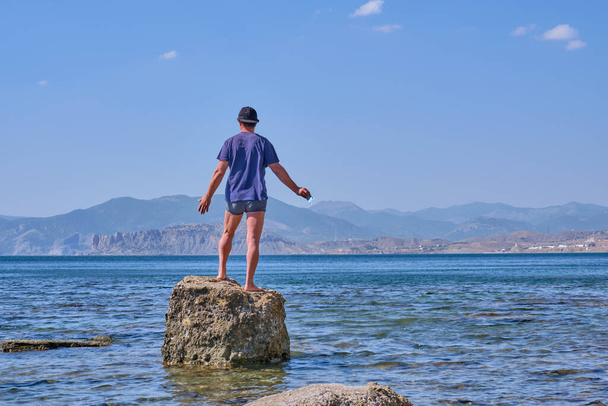 Ανώτερος Ασιάτης με σορτς, t-shirt και καπέλο του μπέιζμπολ, κρατώντας τη μάσκα του προσώπου του, στέκεται μόνος του σε μια πέτρα σε ένα κόλπο στη θάλασσα πάνω από ένα φόντο βουνών - Φωτογραφία, εικόνα