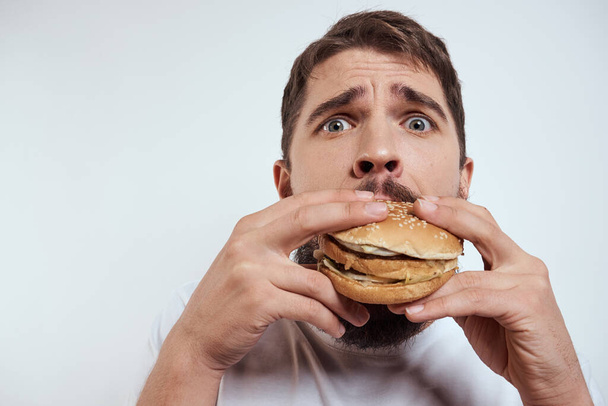 Чоловік їсть гамбургер на світлому тлі в білій футболці обрізаний вид крупним планом голодний фаст-фуд
 - Фото, зображення