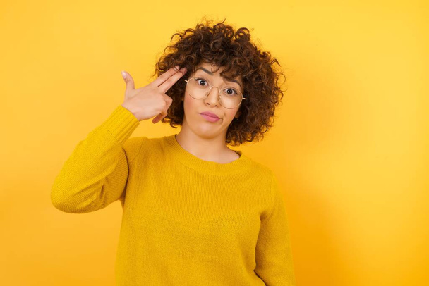 Unhappy woman in yellow sweater with curly hair in studio, imitates gun shoot makes suicide gesture keeps two fingers on temples has puzzled expression dressed in casual clothes stands indoors. - Photo, Image
