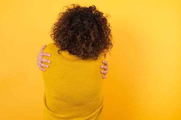 πίσω όψη της γυναίκας με σγουρά μαλλιά φορώντας κίτρινο πουλόβερ αγκαλιάζει τον εαυτό της, Αυτο αγάπη και την αυτοφροντίδα - Φωτογραφία, εικόνα