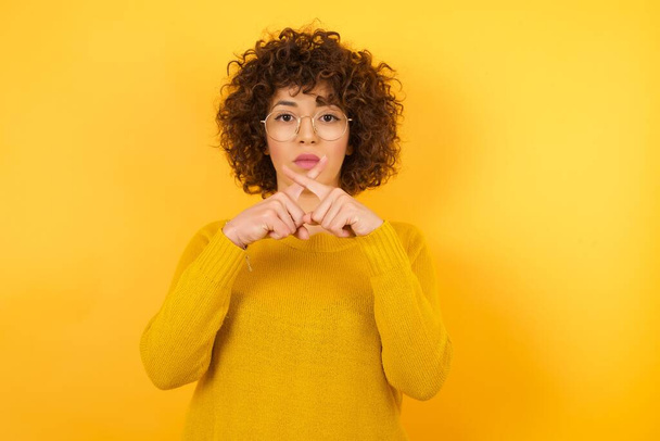 Curly Hair, кавказька жінка в жовтій студії, сердитий вираз перетинає пальці роблячи негативний знак. - Фото, зображення