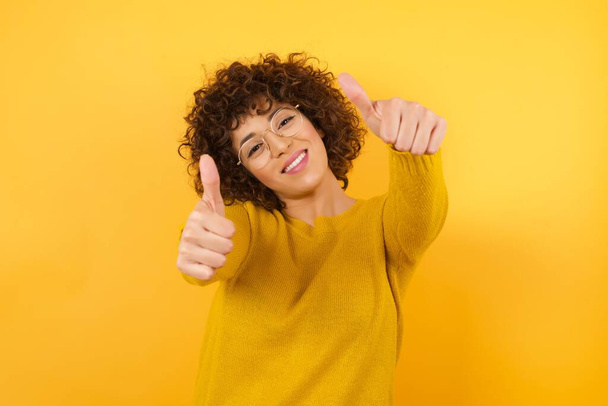 smiling woman with curly hair in yellow studio, approving doing positive gesture with hands, thumbs up smiling and happy for success. Winner gesture. - Photo, Image