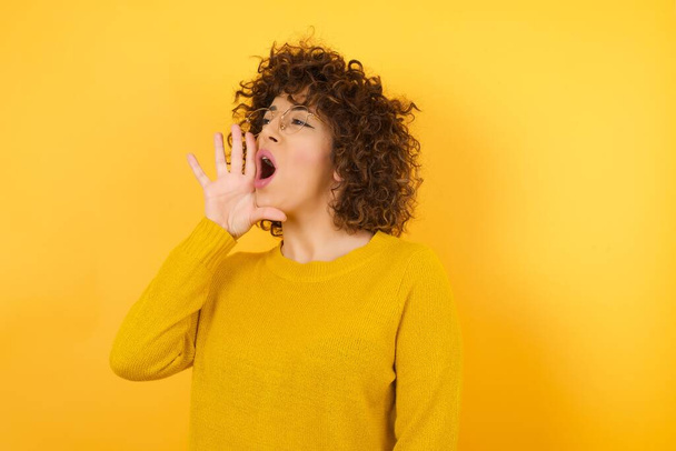 woman in yellow sweater with curly hair in studio, shouting and screaming loud to side with hand on mouth. scream with hand at mouth, opened mouth  - Фото, изображение