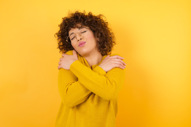 woman in yellow sweater with curly hair in studio, Hugging herself happy and positive, smiling confident. Self love and self care - Fotó, kép