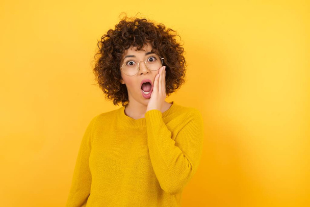 mujer sorprendida con el pelo rizado en estudio amarillo, cara asombrada con la boca abierta  - Foto, imagen