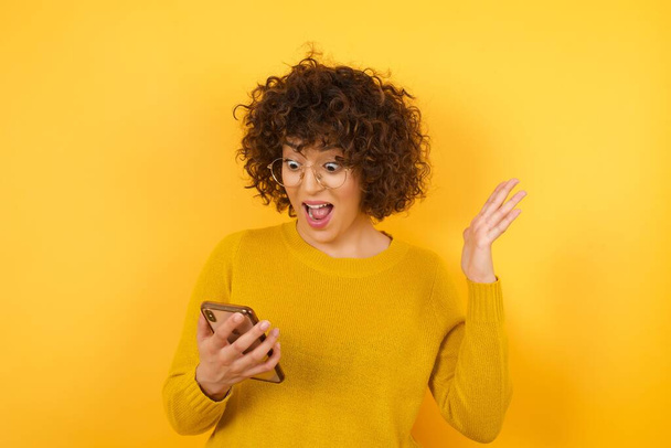 έκπληξη γυναίκα με σγουρά μαλλιά σε κίτρινο στούντιο, κρατώντας και περιήγηση στο κινητό τηλέφωνο  - Φωτογραφία, εικόνα