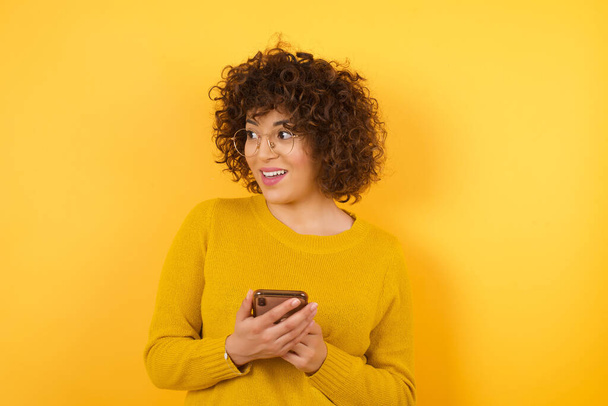 щаслива жінка з кучерявим волоссям в студії з смартфоном, озираючись, копіює простір стіни
 - Фото, зображення