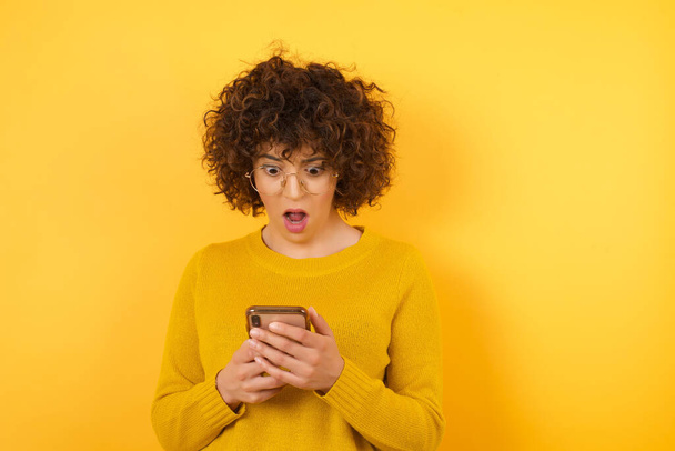 здивована жінка з кучерявим волоссям в жовтій студії, тримає і переглядає мобільний телефон
  - Фото, зображення