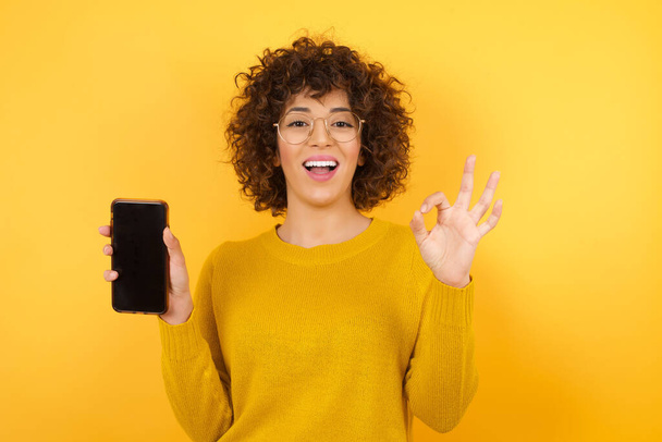 femme heureuse avec les cheveux bouclés en studio avec smartphone, montrant ok signe  - Photo, image