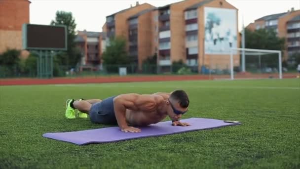 Izmos férfi napszemüvegben edz a városi stadionban, és kihívást jelentő fekvőtámaszokat csinál a szőnyegen. Közelkép. Homályos háttér - Felvétel, videó