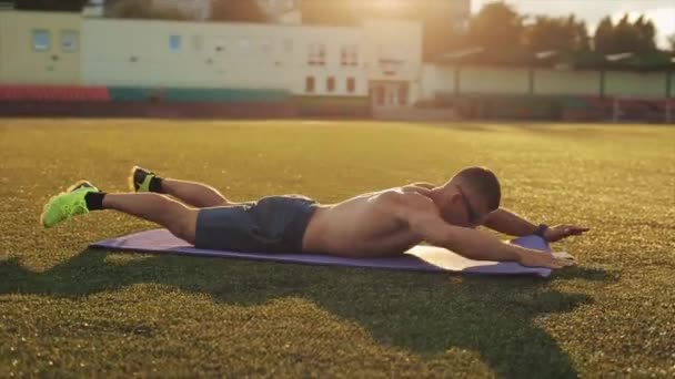 Egy kék rövidnadrágos testépítő edz a stadionban, és ugrál és fekvőtámaszt készít a napos égbolt hátterében. Közelkép. Oldalnézet - Felvétel, videó