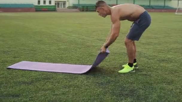 Homem atlético de calções azuis dobra o tapete esportivo depois de treinar no estádio da cidade. Vista lateral. Close-up - Filmagem, Vídeo