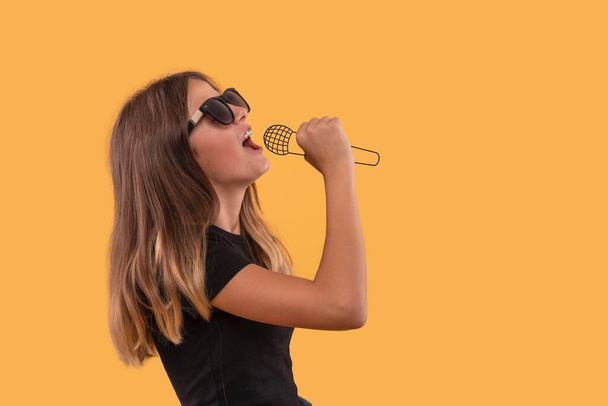 Studio shot of young girl dreaming of a musical career singing into an imaginary microphone,  against yellow background with copy space  - Photo, Image