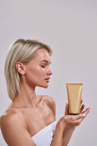 Portrait of young woman with perfect glowing skin looking at sunscreen, bronzer, skincare product while holding and advertising it, posing isolated over grey background - Fotoğraf, Görsel